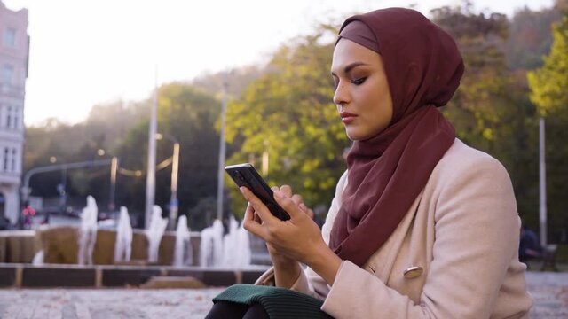 A young beautiful Muslim woman swipes on a smartphone in a street in an urban area - a fountain in the blurry background - closeup