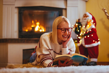 Fototapeta na wymiar Relaxed woman lying on sofa with book in hand next to Christmas tree. Christmas Reading. Young beautiful girl is reading a book next to the fireplace and the Christmas tree. Christmas holidays
