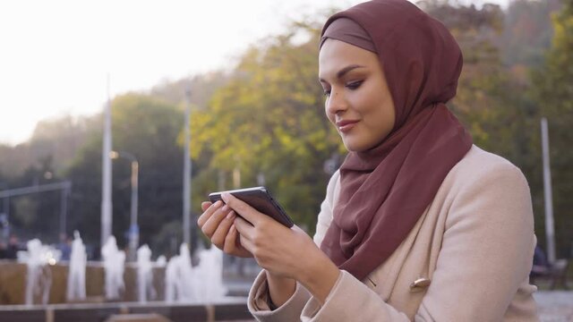 A young beautiful Muslim woman looks at a smartphone with a smile in a street in an urban area - a fountain in the blurry background - closeup