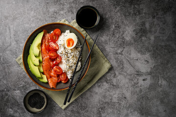 Poke bowl - green bowl with white rice, smoked salmon, cherry tomatoes and avocado, sesame seeds, soy sauce and chopsticks on a grey concrete surface. Top view, negative space