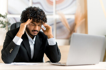 Exhausted tired young adult curly-haired Indian businessman, broker or manager sits at the table in the office, massaging temples, he needs rest, is stressed, headache, migraine. Feelings unwell