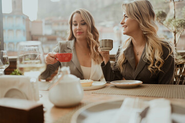 Close-up portrait of two female friends in strict suits laughing drinking coffee and wine on terrace outside at summer street cafe on background buildings Old Tbilisi city, Georgia. High quality photo