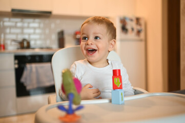 Little happy cute baby toddler boy blonde sitting on baby chair playing with constructor. Baby facial expressions indoors at home kitchen interior with toys. Healthy happy family childhood concept