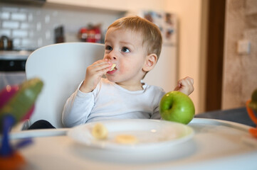 Little happy cute baby toddler boy blonde sitting on baby chair playing with apple. Baby facial expressions indoors at home kitchen interior with food. Healthy eating happy family childhood concept