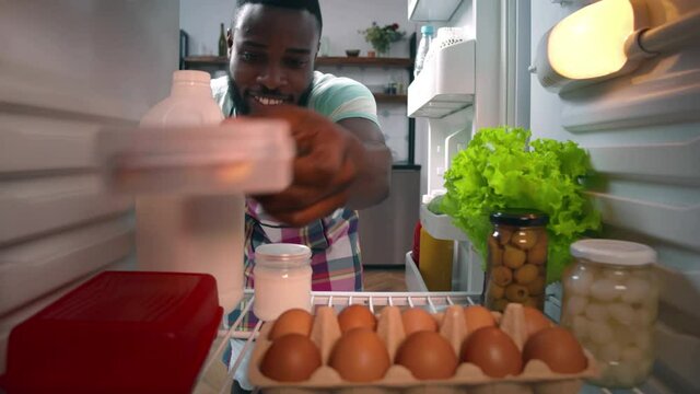 Young African-American ethnicity man in apron taking food from fridge to cook dinner. Realtime