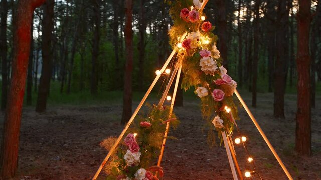 Bohemian tipi wooden arch decorated with burning candles, roses and pampass grass, wrapped in fairy lights illumination on outdoor wedding ceremony venue in pine forest at night. Bulbs garland shines.