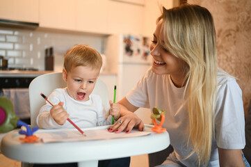 Obraz na płótnie Canvas Little cute baby toddler boy blonde sitting on baby chair learning to draw. Beautiful young mom and son playing spend time together indoors at home with toys. Healthy happy family childhood concept