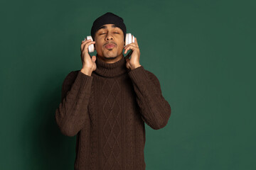 Young handsome man in warm clothes and hat listening to music isolated on green color studio...