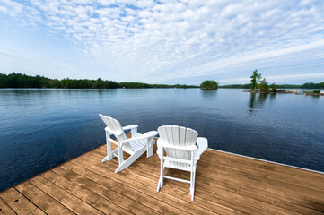 Wooden dock with two white Adirondack chairs facing the blue water of a lake in Muskoka Ontario,...