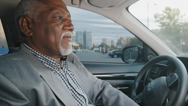 Old african american man driving car serious pensive elderly male leaves parking lot mature businessman looking closely at road drives up approaches intersection experienced driver ride on highway