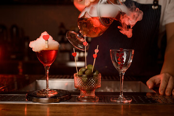 great view on glass of cocktail with foam and vase with olives and glass into which the bartender pours steaming beverage