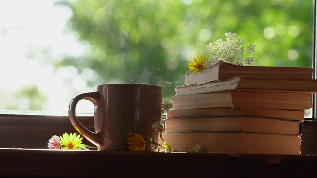 Close-up view 4k stock video footage of brown mug with hot steaming morning drink standing on windowsill of rural cottage, near laying stack of old paper books with cute small meadow flowers