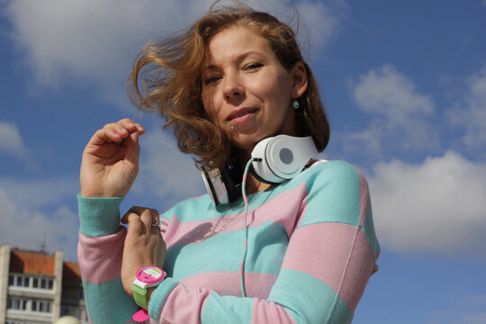 Portrait of a woman in a striped sweater of pastel colors with white headphones against a background of clouds and sky