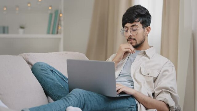Young guy arab freelancer business man male bearded student in glasses lies on couch at home with laptop working studying reading online thinking about connection problem rubs chin pondering solution