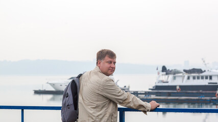 A man with a backpack in a wet jacket is waiting for his ship in rain.