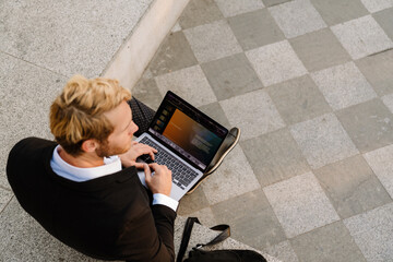 Obraz premium Blonde man working with laptop while sitting on stone bench
