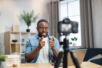 Positive young guy recording video blog on camera while staying at home. African american man in eyewear and casual outfit creating new content for his social networks. - obrazy, fototapety, plakaty