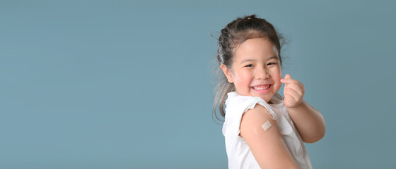 Coronavirus Vaccination Advertisement. Happy Vaccinated Little asian girl Showing Arm With Plaster Bandage After Covid-19 Vaccine Injection Posing Over Blue Background, Smiling To Camera. 