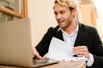 Blonde man working with laptop and papers in cafe