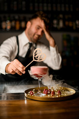 hand of bartender holds wooden decoration over wine glass with smoking cocktail on tray with strawberries on the bar counter.