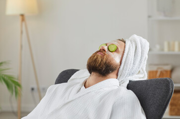 Adult man in bathrobe and towel turban enjoying spa day at home and relaxing in comfortable chair or armchair with facial beauty mask on face and fresh cucumber slices on eyes. Skin care concept
