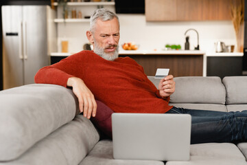 Internet theft blackmailing fraud online. Doubting caucasian mature man paying with credit card online, doing shopping , e-banking, e-commerce at home