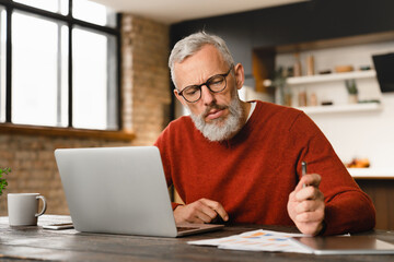 Concentrated mature middle-aged man businessman freelancer tutor doing paperwork, working remotely on laptop, watching webinars, e-learning online at home