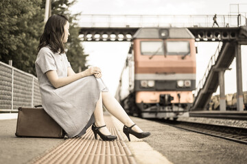 A beautiful girl on the platform is sitting on a suitcase against the background of an approaching train. Vintage style processing. The concept of traveling by train, rail transport.