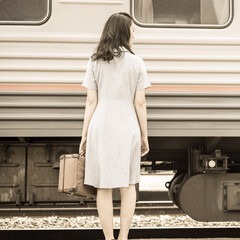 A young woman with a suitcase is standing with her back against the background of a moving train. Retro style. Concept: departing train, departure, arrival.