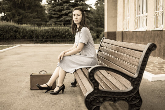 A young woman in retro style with a vintage suitcase is sitting on a bench on the platform waiting for her train.