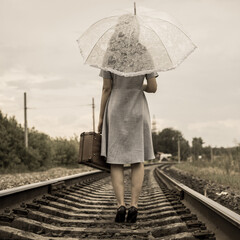 Vintage style. A girl in a dress and heels with an umbrella and a suitcase goes by rail on foot. Rear view. No face. A retro-style girl walking off into the distance by rail.