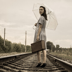 A young woman with an umbrella and a suitcase walks away along the rails of the railway and looks...