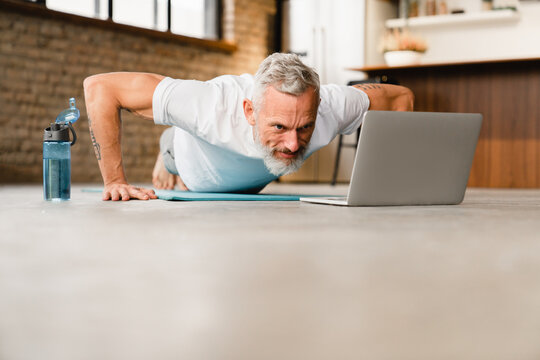Handsome fit slim man doing push-ups at home on fitness mat, watching online video tutorial training, vlogging and blogging his exercises at home