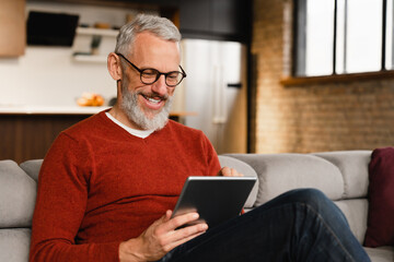 Smiling caucasian middle-aged mature man in glasses using digital tablet for e-learning, paying...