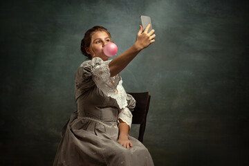 Retro portrait of young romantic girl in gray dress of medieval style using phone isolated on dark...