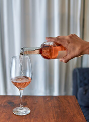 man's hand pouring rose wine into a glass on a wooden table with a white curtain in the background at home