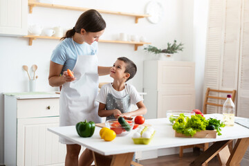 Cheerful mother proud her self-dependent son. Child unaided is cooking salad at kitchen. Healthy food concept.