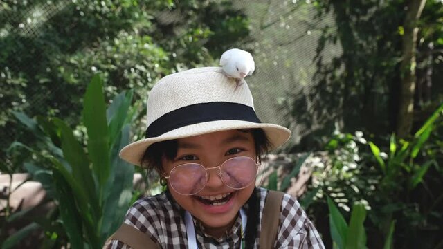 Asian girl in a brown overalls with a hat is feeding a bird in her hand up close.