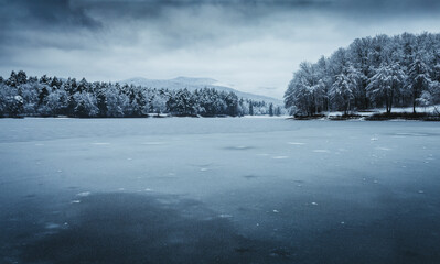 Dark landscape photo of frozen and snowy lake with forest and big hills on background - winter time. Frozen and cold lake with cloudy and dramatic sky - blue tones.