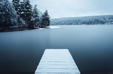 Dark landscape photo of mole (pier) covered of snow with frozen lake on background - winter time. Frozen and cold lake with hills and forest on background - blue tones.