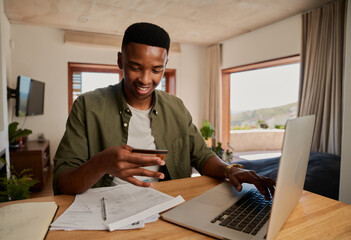 Fototapeta premium Happy young adult black male smiling, making online payment on laptop in modern apartment. 