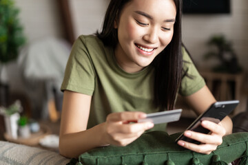 Fototapeta na wymiar Online shopping. Young asian woman sitting at home and making purchase on mobile phone app, holding credit card