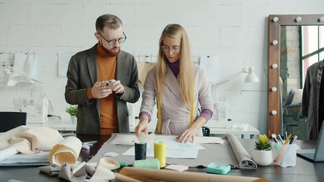 Clothes designers man and woman are looking at drawings choosing fabric and using smartphone indoors in workshop. People and occupation concept.