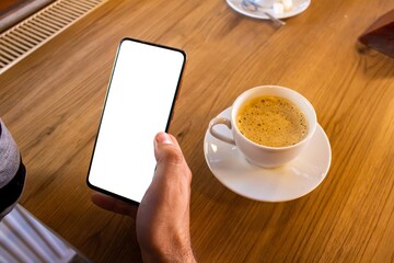 Hand holding mobile phone with blank white screen while drinking coffee.
