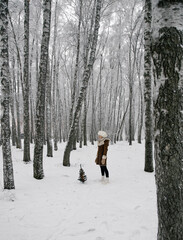 A girl in a sheepskin coat, mittens, white boots and a hat with earflaps in a winter snowy forest stands in a birch forest with an artificial little decorated Christmas tree