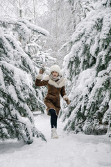 Vertical photo of a young European girl in a sheepskin coat, mittens, a white hat with earflaps and boots, walking among a pine forest in the snow in winter