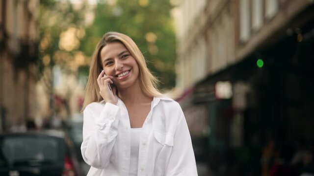 Cheerful woman talking on the phone in the street wearing a white clothes. Portrait of modern happy casual smiling millennial girl talking phone. Happy young lady talking on mobile phone on the street