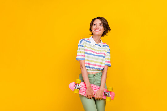 Photo of young girl happy positive smile dream curious look empty space hold skateboard isolated over yellow color background