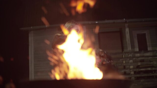 A Caucasian girl in a black jacket warms up near the fire. A European woman stands at the dacha at night and strokes a black dog. Green wooden country house 