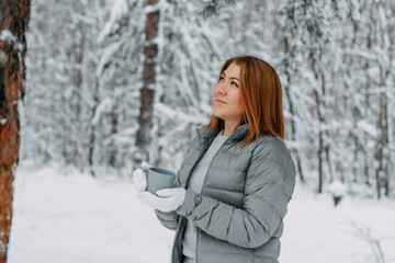 A cute girl with short hair in a gray jacket and knitted white mittens is drinking hot tea in a snowy forest. Cozy and warm in the winter season